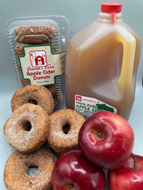 A container of apple cider donuts, a jug of apple cider, and fresh red apples arranged together.