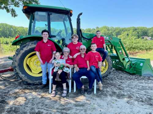 A group of six people in red shirts poses in front of a green tractor on a sunny day in a field.