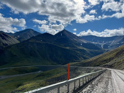 A winding gravel road leads through lush green mountains under a partly cloudy sky.