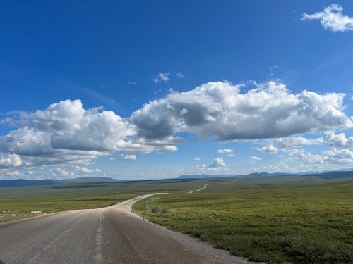 A winding road stretches through a vast green landscape under a blue sky filled with fluffy white clouds.