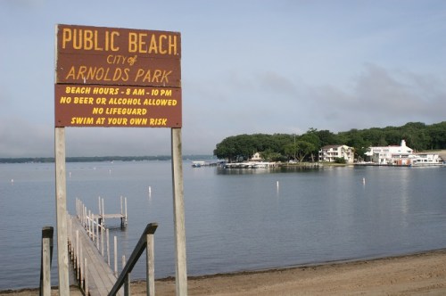 Sign for Public Beach at Arnolds Park, listing hours and rules, with a calm lake and shoreline in the background.