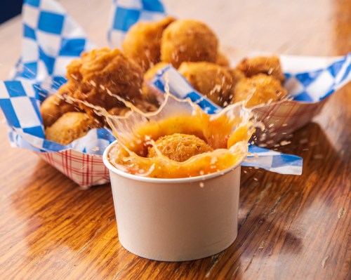A bowl of dipping sauce splashes as fried snacks sit in the background, all on a wooden table with checkered paper.
