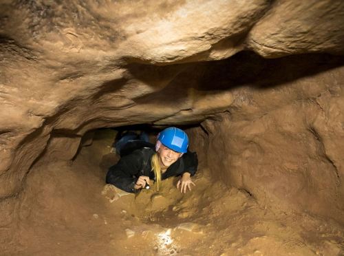 A person in a blue helmet crawls through a narrow cave passage, smiling and holding a flashlight.