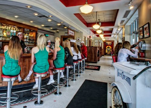 A retro diner interior with patrons at the bar, wearing green outfits, and an ice cream cart in the foreground.