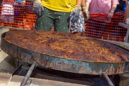 A large, dark-brown dish of cooked food on a grill, with people in the background watching.