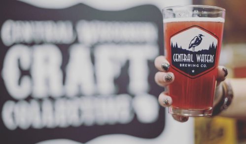 A hand holds a glass of reddish beer with a logo for Central Waters Brewing Co. in front of a blurred background.