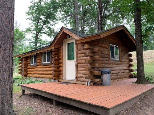 A rustic log cabin with a wooden deck, surrounded by trees and greenery.