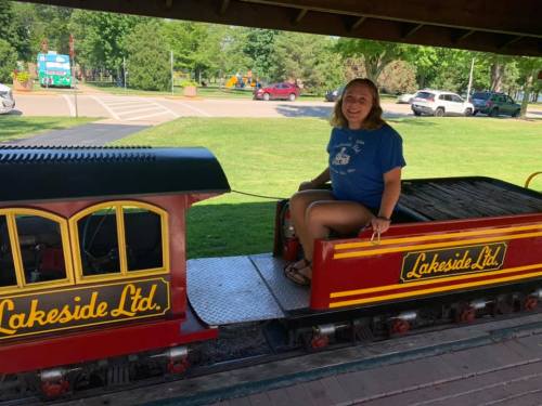 A girl sits on a colorful miniature train in a park, surrounded by green grass and trees.