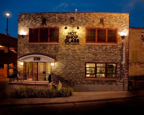 Exterior of a brick building at night, featuring a sign that reads "Hot Shop Glass" and illuminated windows.