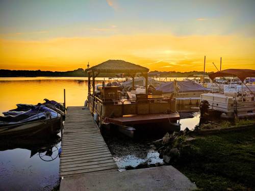 A serene sunset over a lake, with boats docked and a wooden pier leading to a gazebo.