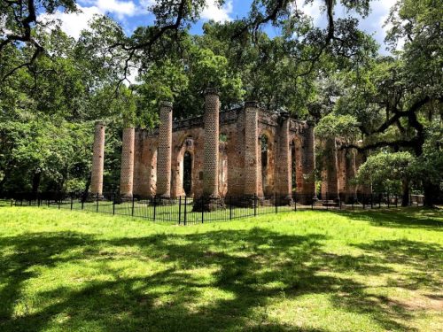 Ruins of a brick structure surrounded by lush greenery and trees, enclosed by a black fence under a blue sky.