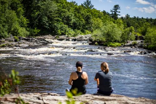 Two people sit by a river, enjoying the view of flowing water and lush greenery under a clear blue sky.