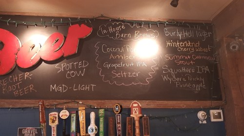 Chalkboard menu listing various beers, including IPAs, stouts, and seltzers, with decorative lights in the background.