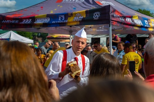 A man in a white shirt and paper hat holds a large sandwich, surrounded by a crowd at an outdoor event.
