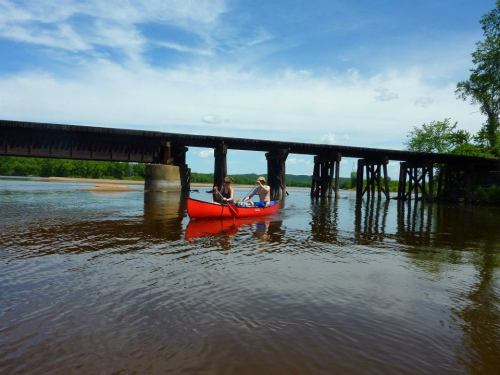 Two people paddle a bright orange kayak under a wooden bridge on a calm river, surrounded by greenery and blue skies.