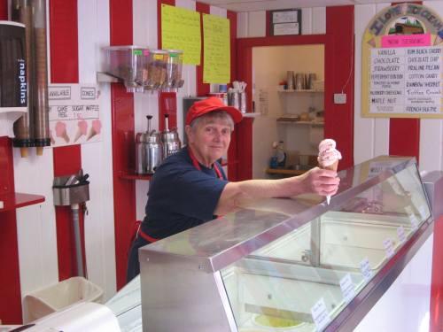 A smiling ice cream shop employee holds a cone with pink ice cream, standing behind a glass display of flavors.