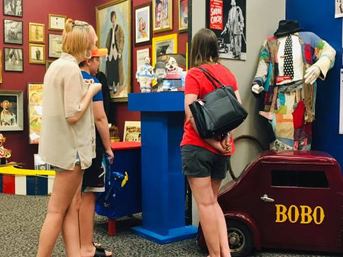 Three visitors observe clown memorabilia in a colorful exhibit, featuring a mannequin and various framed photos.