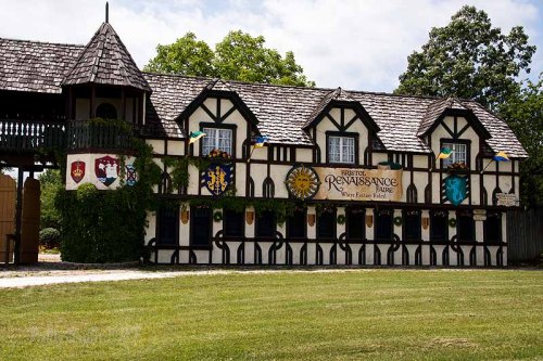A charming Tudor-style building with banners and shields, set in a grassy area, evoking a Renaissance fair atmosphere.