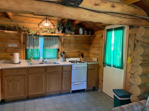 Cozy log cabin kitchen with wooden cabinets, green curtains, and a gas stove, featuring rustic decor and natural light.