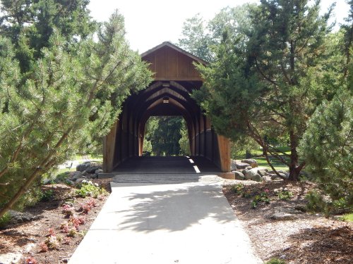 A wooden covered bridge surrounded by trees and landscaped gardens, leading to a pathway.