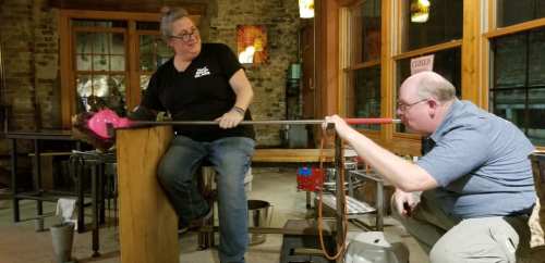 A woman demonstrates glassblowing techniques to a man in a workshop, with tools and glass materials around them.