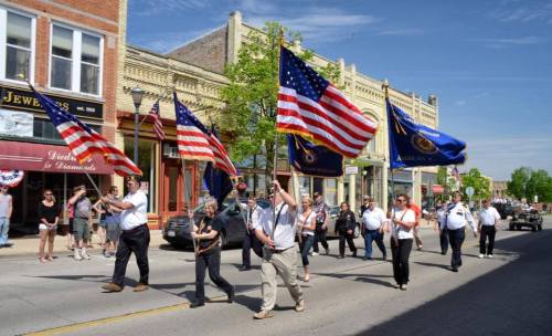 A parade featuring people carrying American flags and banners, with spectators lining the street on a sunny day.