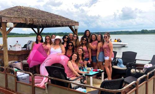 A group of women in matching pink tops enjoy a lakeside gathering on a boat, with drinks and a pink float nearby.