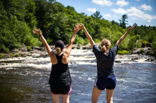Two women stand by a river, raising their arms in celebration against a backdrop of trees and a blue sky.