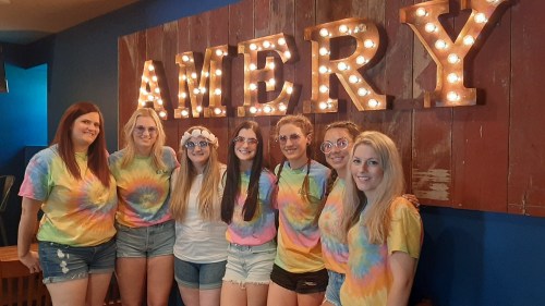 A group of seven women in tie-dye shirts and colorful glasses pose together in front of a lit sign that says "AMERY."
