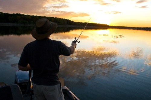 A person fishing from a boat at sunset, casting a line over calm waters reflecting the colorful sky.