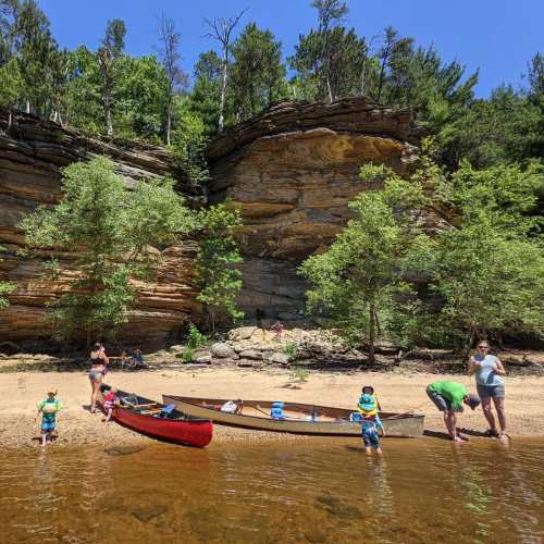 A family enjoys a sunny day by the river, with children playing near a canoe on the sandy shore and cliffs in the background.