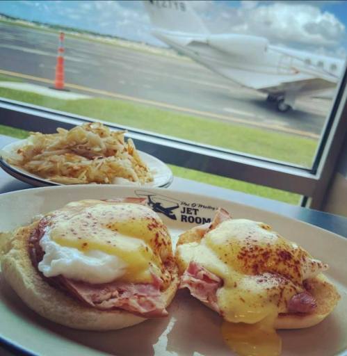 A plate of eggs Benedict with ham, served with hash browns, and a view of a small airplane outside the window.