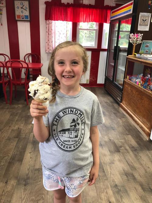 A smiling girl in a gray t-shirt holds a large ice cream cone in a colorful ice cream shop.