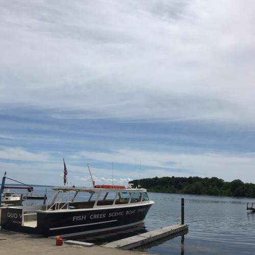 A boat named "Fish Creek Scenic Boat Tours" docked at a pier on a calm lake with trees in the background.