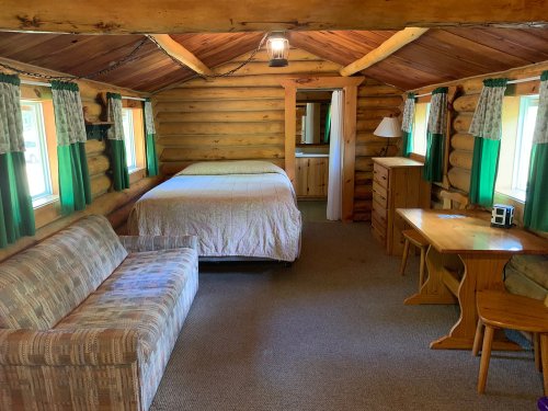 Cozy log cabin interior featuring a bed, sofa, table, and windows with green curtains. Warm, rustic atmosphere.
