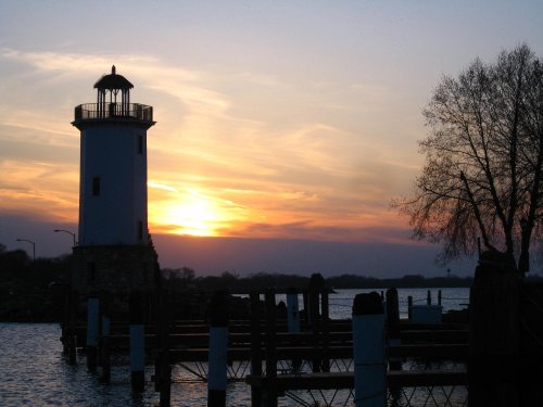 Silhouette of a lighthouse at sunset, with a calm water surface and trees in the background.