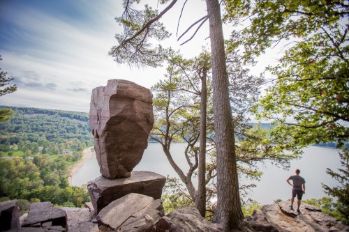 A person stands near a large rock formation overlooking a serene lake surrounded by trees and hills.