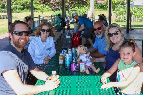 A group of six people sitting at a picnic table, smiling and enjoying a sunny day outdoors.