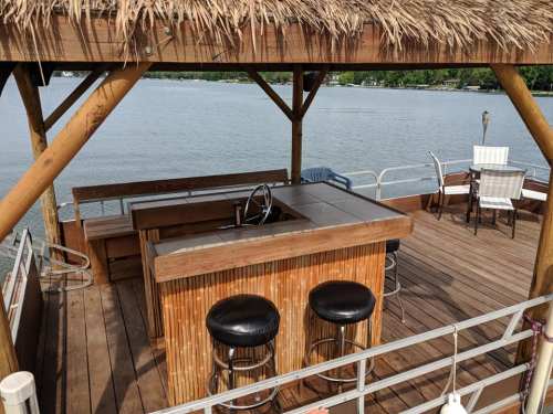 A wooden deck with a tiki-style bar, stools, and a table overlooking a calm lake.