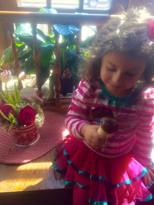 A young girl in a striped shirt and pink skirt enjoys an ice cream cone, surrounded by flowers and plants indoors.