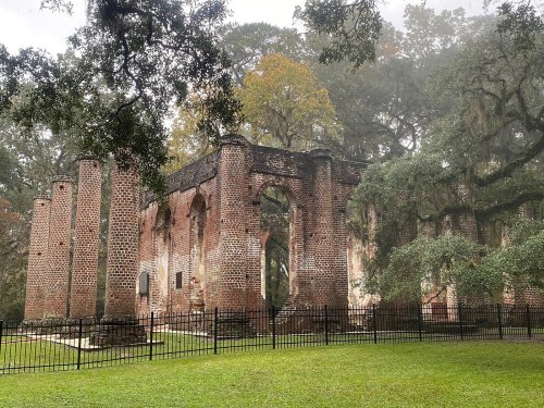 Ruins of a brick structure surrounded by trees and mist, with a green lawn and a black fence in the foreground.