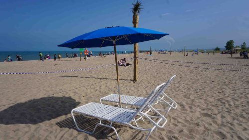 Two white lounge chairs under a blue umbrella on a sandy beach, with people enjoying the sun in the background.