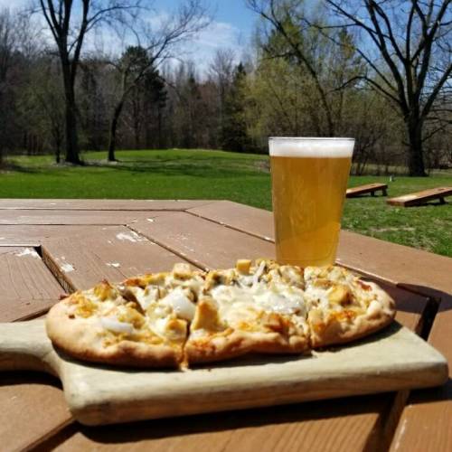 A wooden table with a pizza on a cutting board and a glass of beer, set against a sunny outdoor park backdrop.