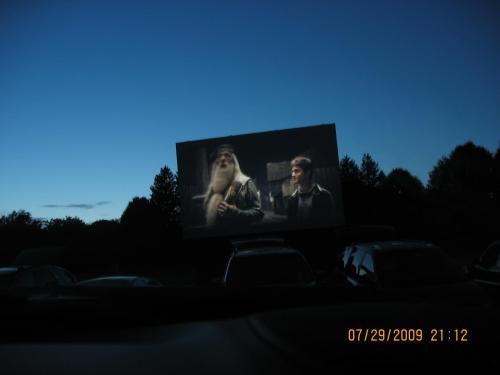 A drive-in movie screen shows two characters in a scene, with a twilight sky and parked cars in the foreground.