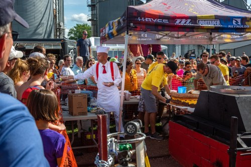 A crowd gathers at an outdoor event as a chef in a white outfit serves cake from a large table.