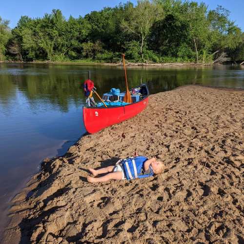A child in a striped shirt lies on a sandy riverbank beside a red canoe, surrounded by trees and calm water.