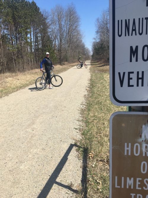 Two cyclists on a gravel path beside a sign warning against unauthorized vehicles, surrounded by trees.