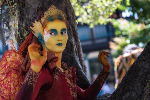 A person with vibrant face paint and a colorful costume poses near a tree, showcasing artistic makeup and elaborate attire.