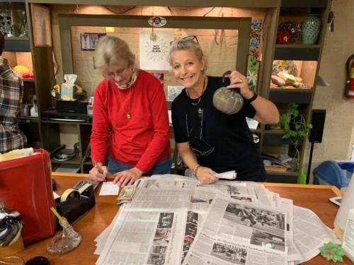 Two women are working at a table covered with newspapers, one writing notes and the other holding a teapot.