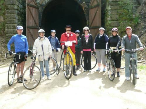 A group of eight cyclists poses with their bikes near a tunnel entrance, smiling and wearing helmets.